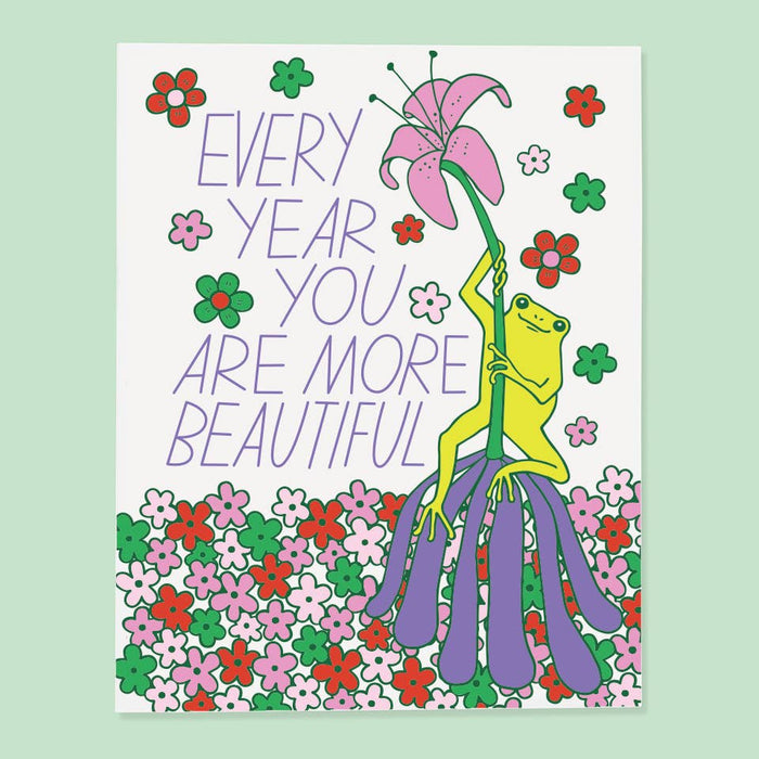 Card with a frog holding a flower and text 'Every year you are more beautiful' on a floral background.