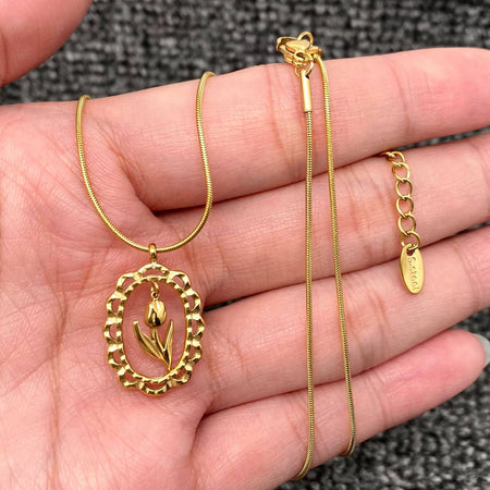 A gold necklace with an oval pendant featuring a tulip design, displayed on a person's hand. The necklace includes a textured beaded border, a fine snake chain, a lobster clasp, and an extender chain with a small brand tag.