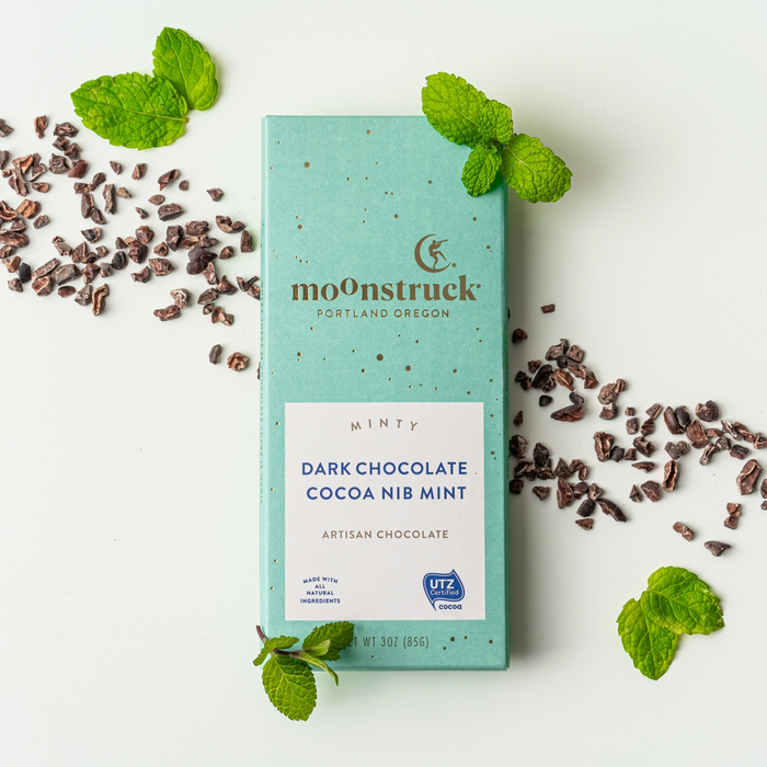 teal moonstruck minty dark chocolate bar on white background with cocoa nibs and mint leaves