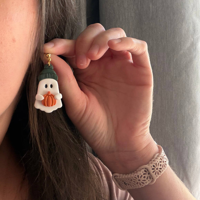 A close-up lifestyle shot of a person holding a small white ghost earring with an orange pumpkin and dark green hat