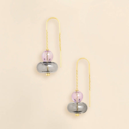 A pair of gold-tone dangle earrings with a translucent lavender purple sphere and a larger smoky gray disc, displayed on a warm cream background.