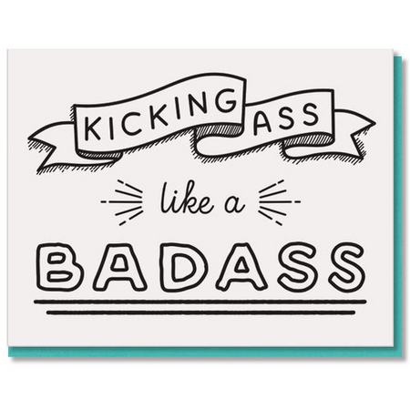 A square greeting card with a white background featuring black hand-drawn text that reads 'KICKING ASS like a BADASS' with decorative ribbon and line elements. A teal card is partially visible behind it on a white background.