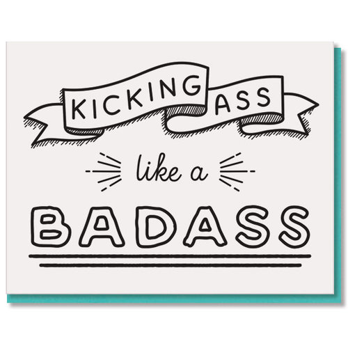 A square greeting card with a white background featuring black hand-drawn text that reads 'KICKING ASS like a BADASS' with decorative ribbon and line elements. A teal card is partially visible behind it on a white background.