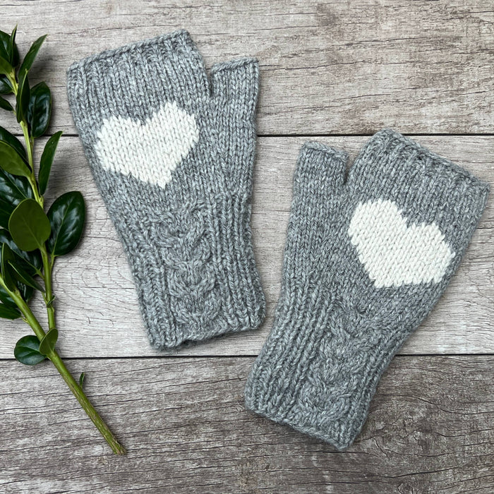 Gray fingerless gloves with heart designs on a wooden surface with a green leafy branch.