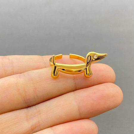 A gold dachshund-shaped ring held on a person's fingers against a soft gray background.