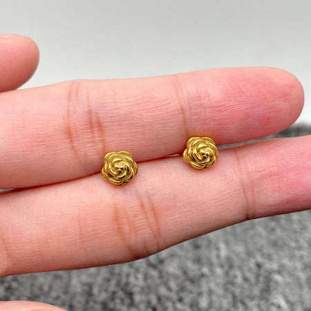 Gold rose-shaped earrings held between two fingers against a neutral background