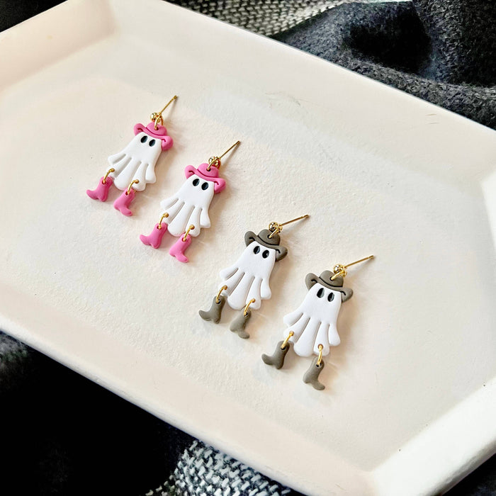 two pairs of dangling ghost earrings displayed on a white ceramic tray. The earrings feature white ghost charms with gold-tone hooks and details: one pair has pink hats and boots, and one pair have brown hats and boots.