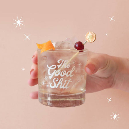 This image features a stylish tumbler designed for those who appreciate a bit of fun and flair in their beverage experience. The tumbler, held in a hand against a soft pink background adorned with sparking stars, is made of glass and boasts a playful screen-printed message that reads "The Good Shit" in a bold, simple font.
