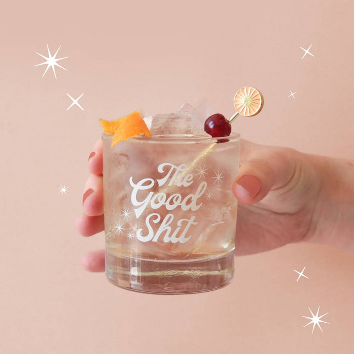 This image features a stylish tumbler designed for those who appreciate a bit of fun and flair in their beverage experience. The tumbler, held in a hand against a soft pink background adorned with sparking stars, is made of glass and boasts a playful screen-printed message that reads "The Good Shit" in a bold, simple font.