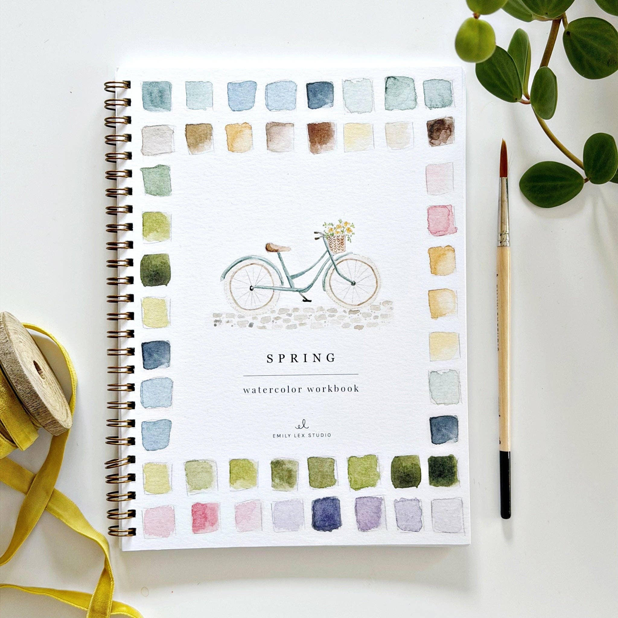 Watercolor workbook with bicycle illustration and color swatches on a white surface with a plant and brush.