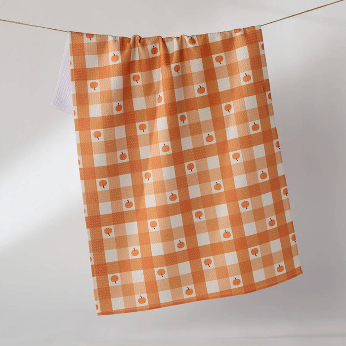 An orange and white gingham kitchen towel with small pumpkin prints, hanging from a string against a light gray background.
