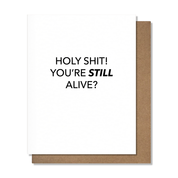 A white greeting card with bold black text reading 'HOLY SHIT! YOU'RE STILL ALIVE?' and a brown kraft envelope behind it, shown on a clean white background.