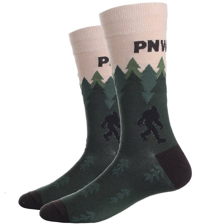 A pair of crew socks featuring a forest pattern with pine trees and Bigfoot silhouettes on a dark green background, with a cream cuff printed with the partial brand name 'PNW'.