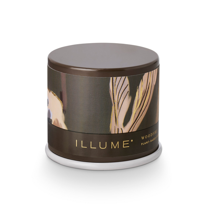 A cylindrical candle tin by ILLUME, featuring a dark brown lid and base with a central label showing a stylized beige and gold leaf pattern on a dark background. The brand name 'ILLUME' is printed in gold on the lower band.