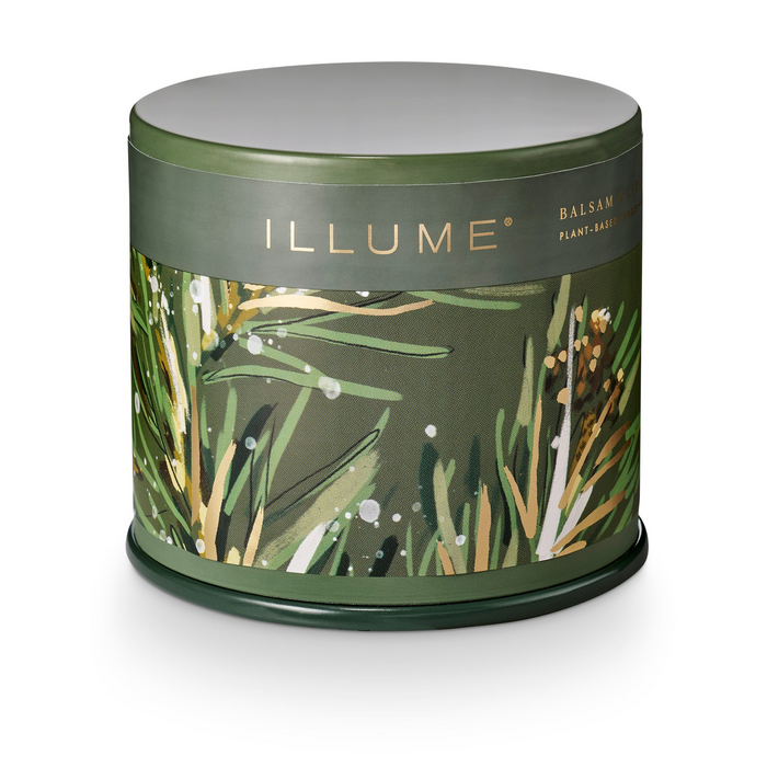 A cylindrical metal tin candle from the brand ILLUME, featuring a dark olive green lid and base with a central label decorated with botanical illustrations of pine needles and leaves in green, gold, and white.