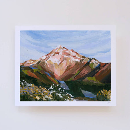 an art print of a mountain landscape with a blue sky, hanging on a white wall. The painting features a mountain with pink and brown tones, green and yellow hills, and a blue lake.