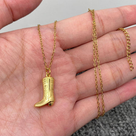 Gold boot-shaped pendant on a chain held in a hand against a neutral background