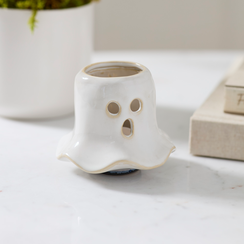 A small white ghost-shaped ceramic planter with a carved face and circular eye cutouts, placed on a light marble surface with a blurred potted plant and books in the background.