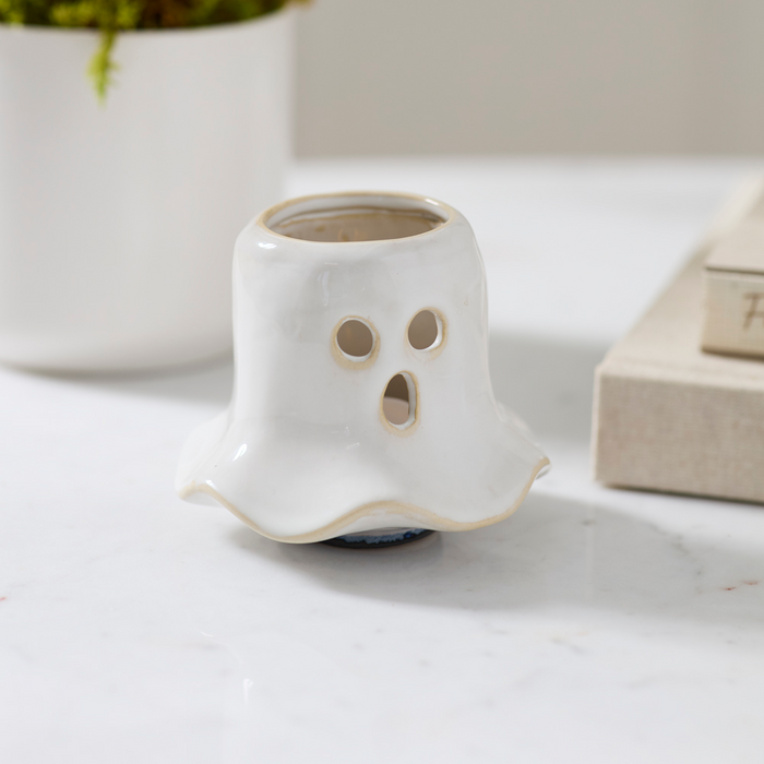 A small white ghost-shaped ceramic planter with a carved face and circular eye cutouts, placed on a light marble surface with a blurred potted plant and books in the background.