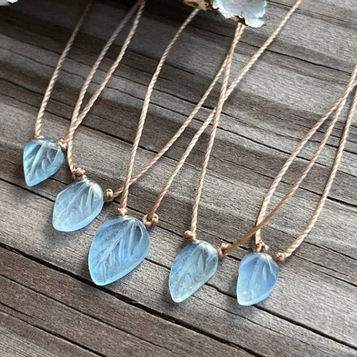 Five leaf-shaped light blue gemstone pendants on thin gold-toned cords, arranged on a weathered wooden surface with dried pale blue flowers in the background.