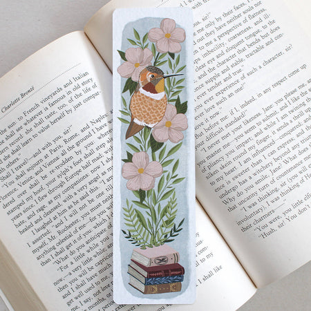 A rectangular illustrated bookmark with a hummingbird, pink flowers, green leaves, and a stack of books, placed on the page of an open book.