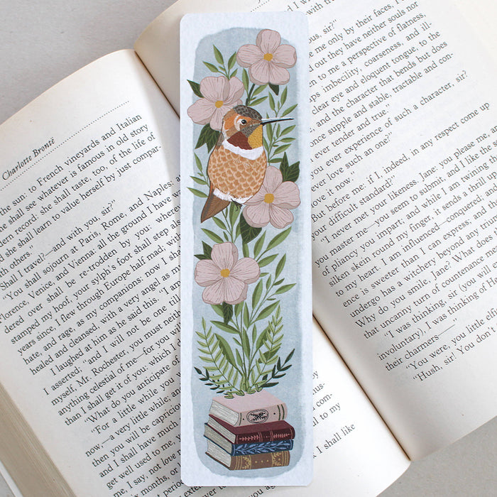 A rectangular illustrated bookmark with a hummingbird, pink flowers, green leaves, and a stack of books, placed on the page of an open book.