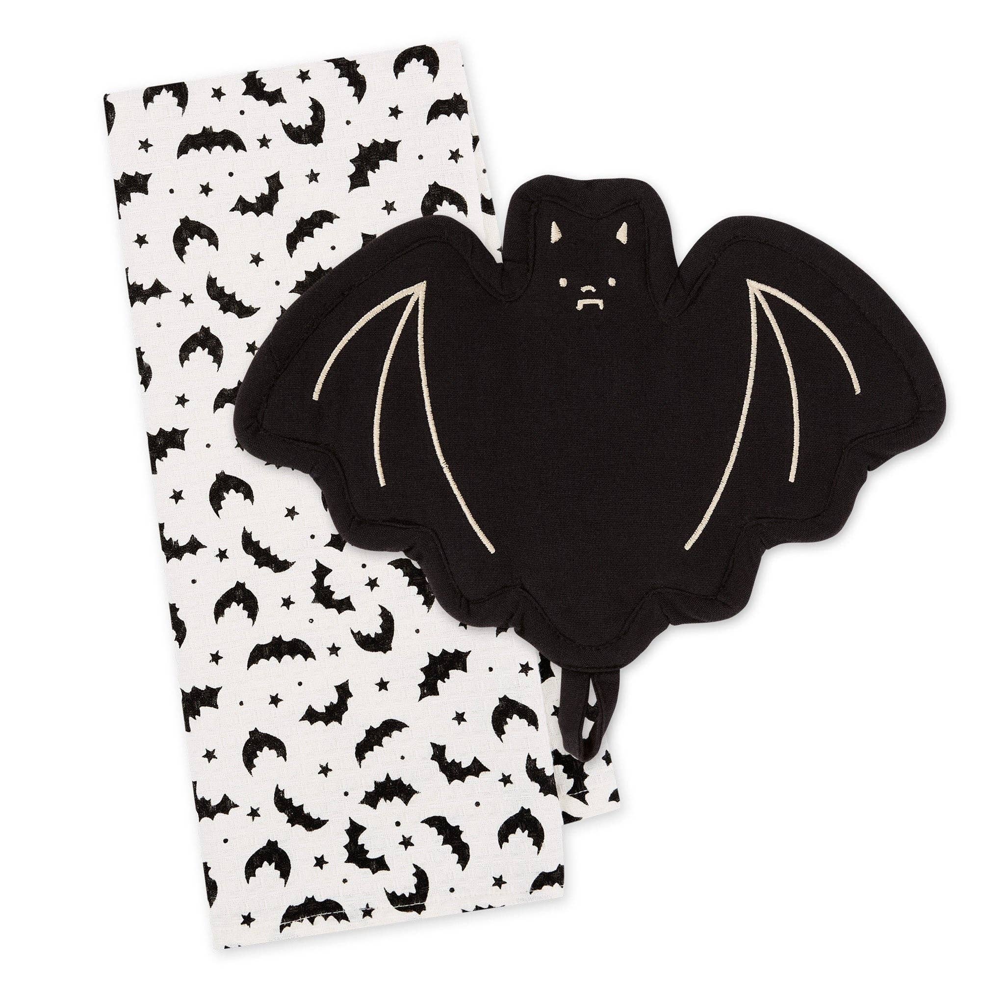 A black bat-shaped potholder with cream-colored stitched wing details, shown next to a white fabric napkin or tea towel printed with a black bat, moon, and star pattern, on a solid white background.