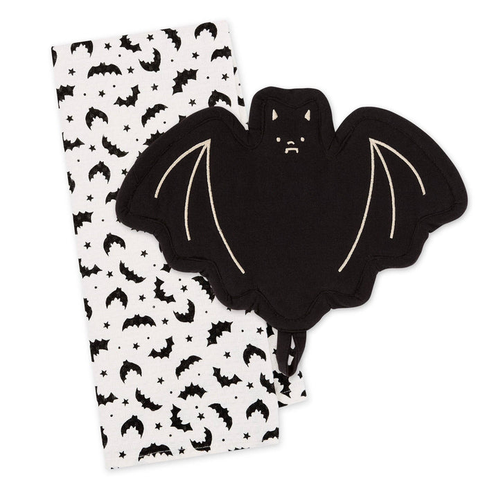 A black bat-shaped potholder with cream-colored stitched wing details, shown next to a white fabric napkin or tea towel printed with a black bat, moon, and star pattern, on a solid white background.