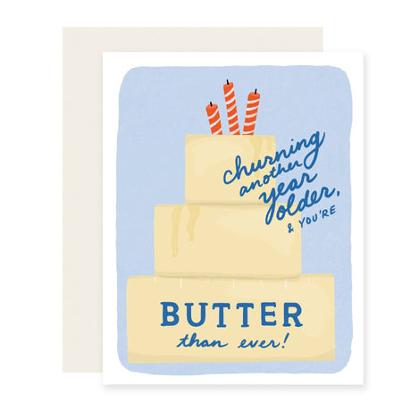A greeting card with a white border and a light blue watercolor-style background, featuring a tiered butter cake with three orange-and-white striped candles and the text 'Churning another year older, & YOU'RE BUTTER than ever!' A plain white envelope is partially visible behind the card.
