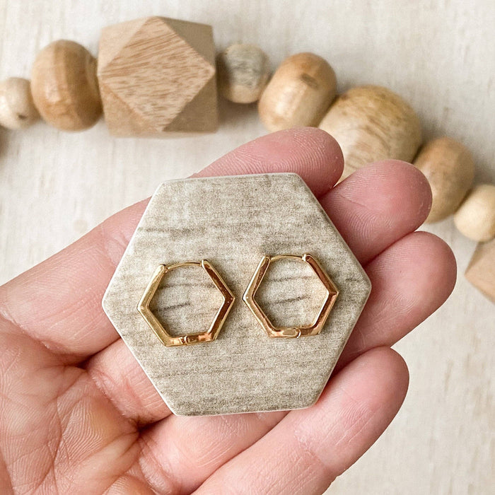 Gold hexagonal earrings on a hexagonal stone display held by a hand with wooden beads in the background.