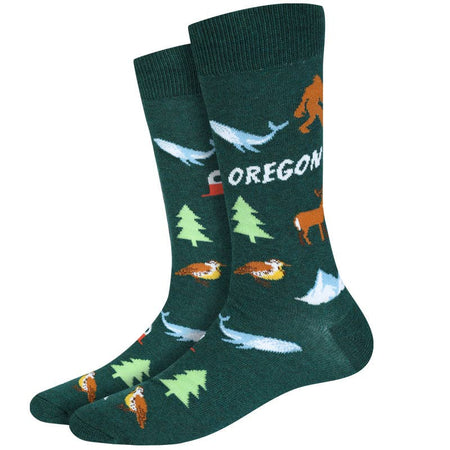 A pair of dark green crew socks with a whimsical Oregon-themed pattern featuring a Bigfoot, blue whales, pine trees, a deer, and the word 'OREGON' in white.