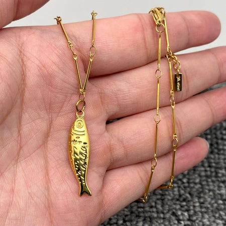 Gold fish-shaped pendant on a chain held in a hand against a neutral background