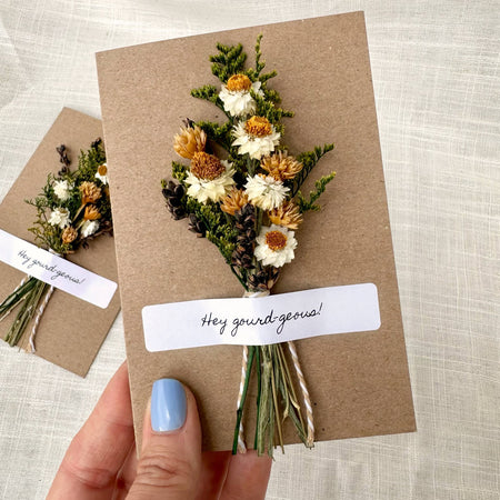 A hand with light blue nail polish holds a brown kraft paper card featuring a pressed dried flower bouquet and a white label that reads 'Hey gourd-geous!'. A second matching card is blurred in the background on a light fabric surface.