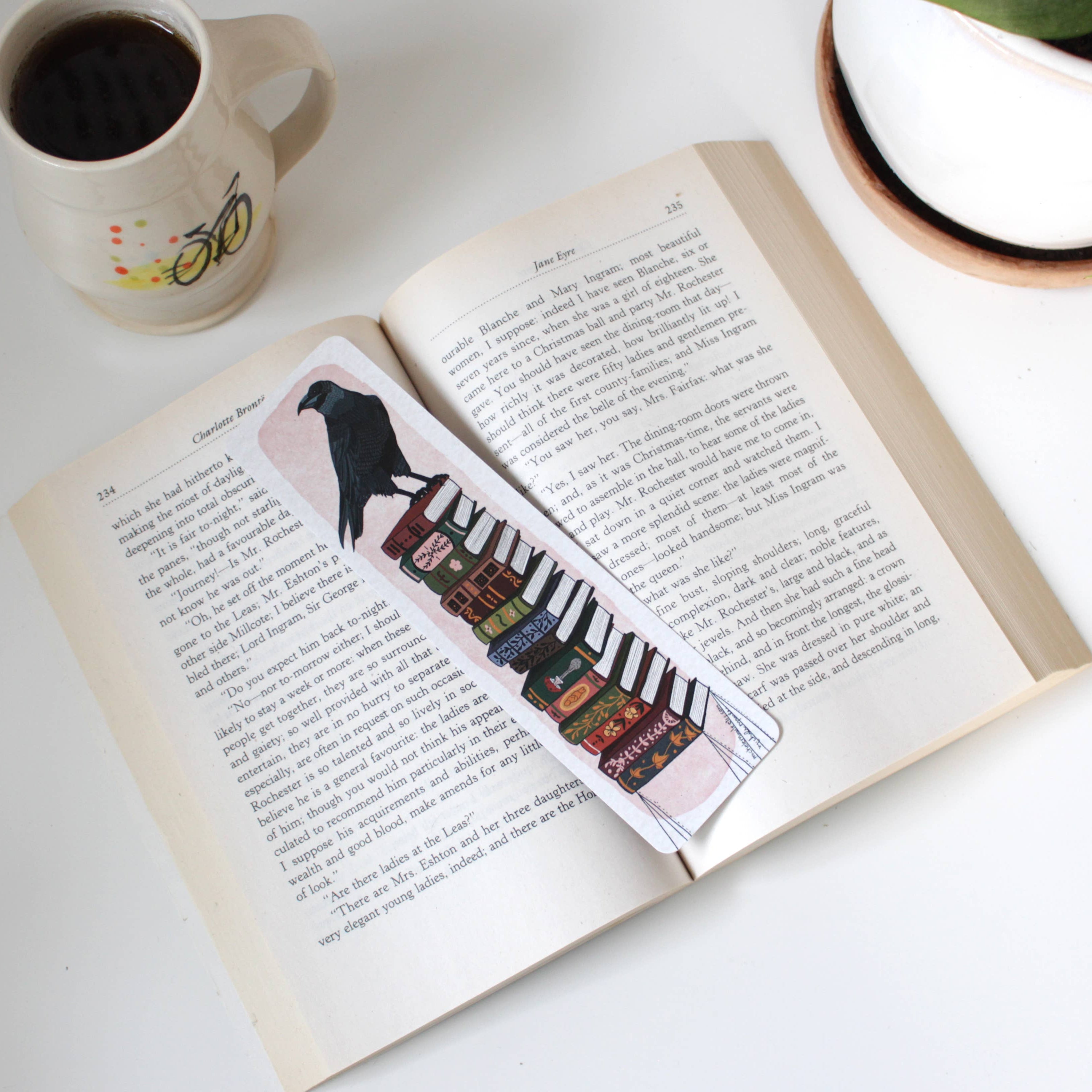 Open book with a bookmark featuring a bird and piano keys, next to a mug on a white surface.