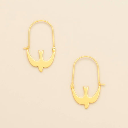 A pair of gold hoop earrings with a small bird silhouette charm, displayed on a light beige background.