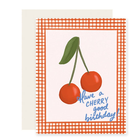 A greeting card with an orange and white gingham border, featuring an illustration of two red cherries with green leaves and the blue handwritten text 'Have a CHERRY good birthday!'. A plain cream envelope is partially visible behind the card.