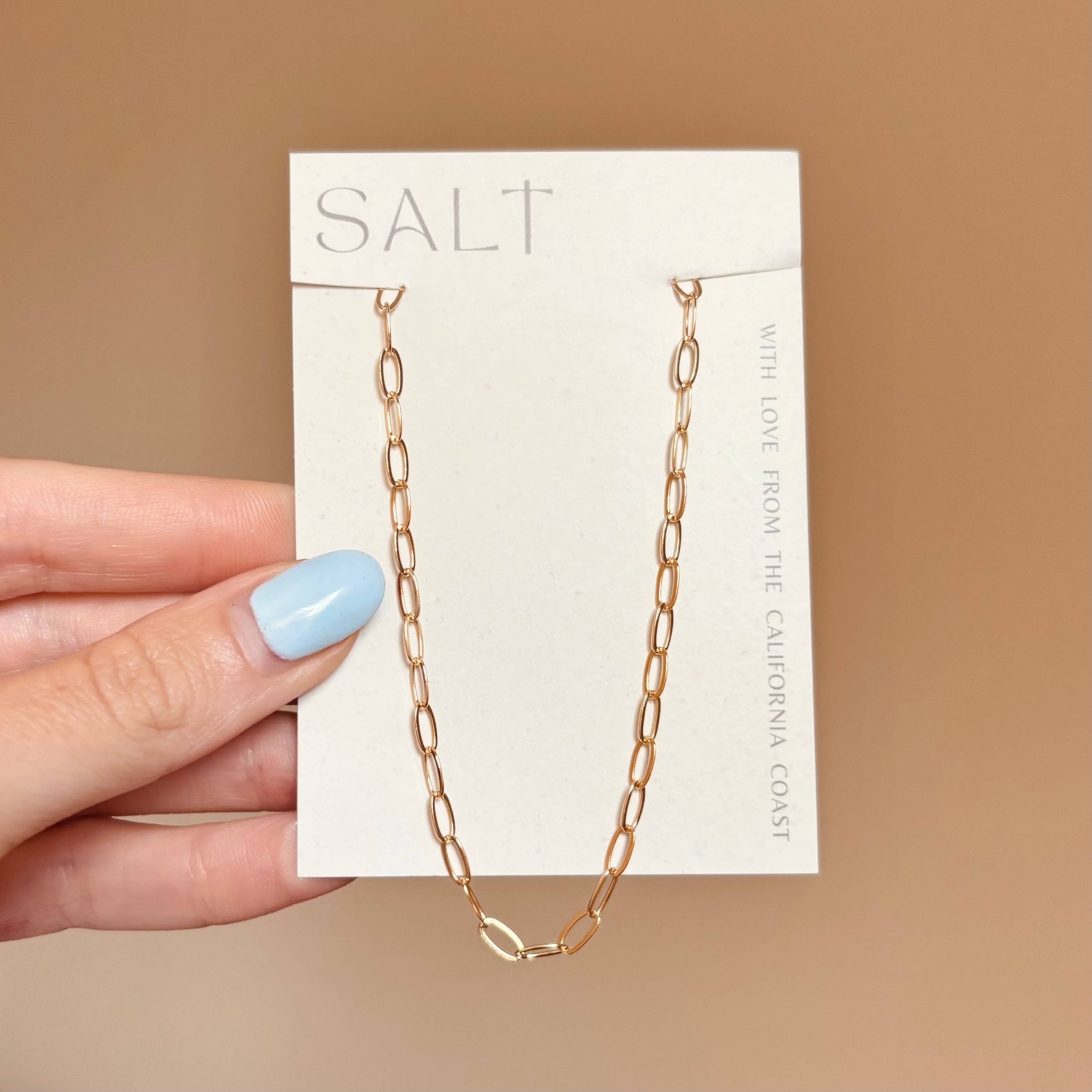 Gold chain necklace on a 'SALT' branded card held by a hand with light blue nail polish.