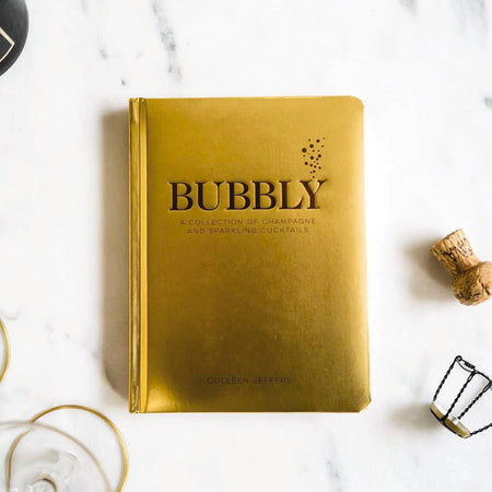 Gold book titled 'Bubbly' on a marble surface with champagne bottle and glasses.