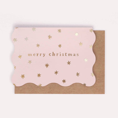 A pink scalloped Christmas greeting card with metallic gold star accents and the words 'merry christmas' in gold foil, shown on top of a brown kraft envelope against a light off-white background.