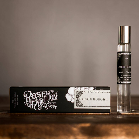 Perfume bottle and packaging with 'Rose & Rebellion Made in the West' branding on a wooden surface.