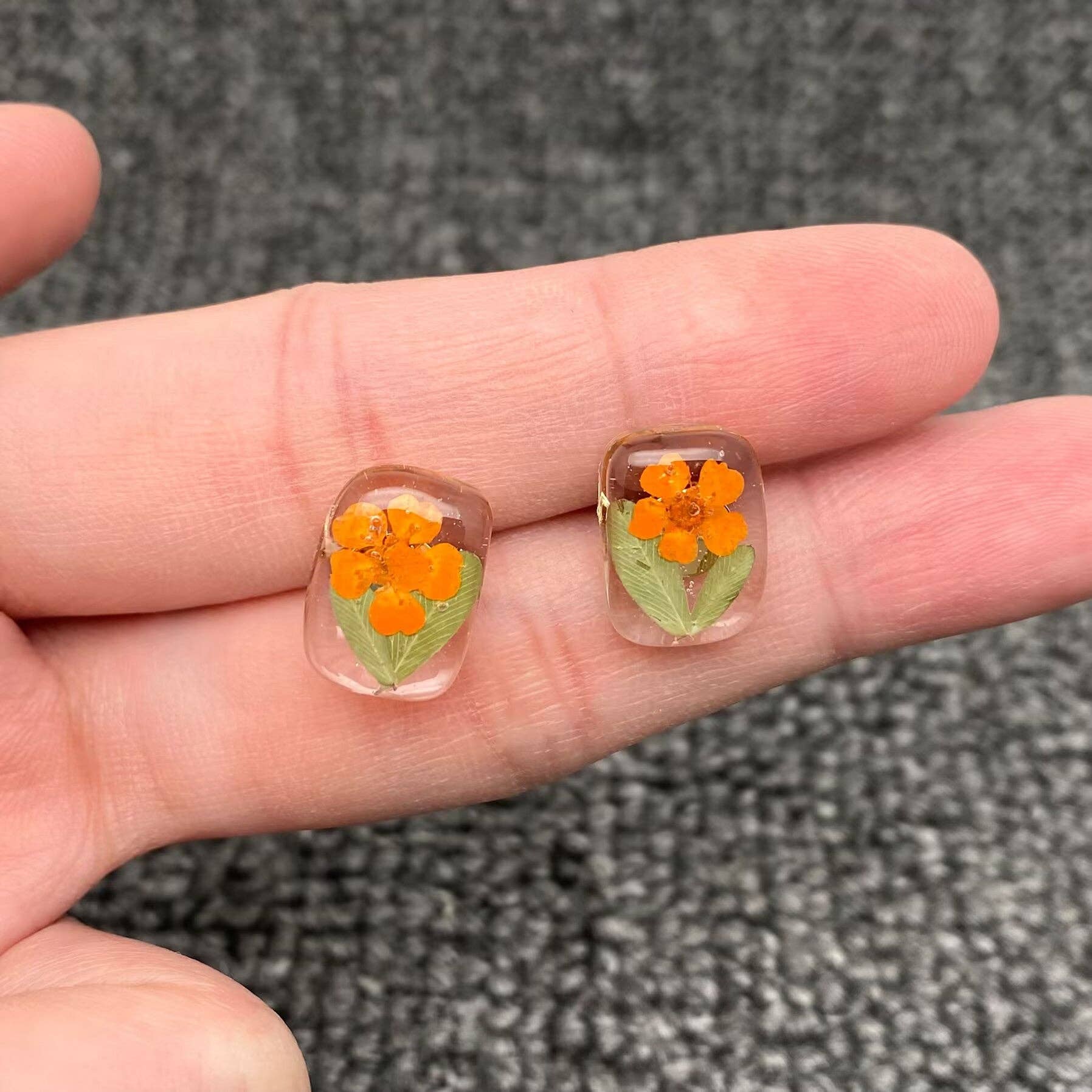Two small transparent earrings with orange floral designs held between fingers on a textured gray background.