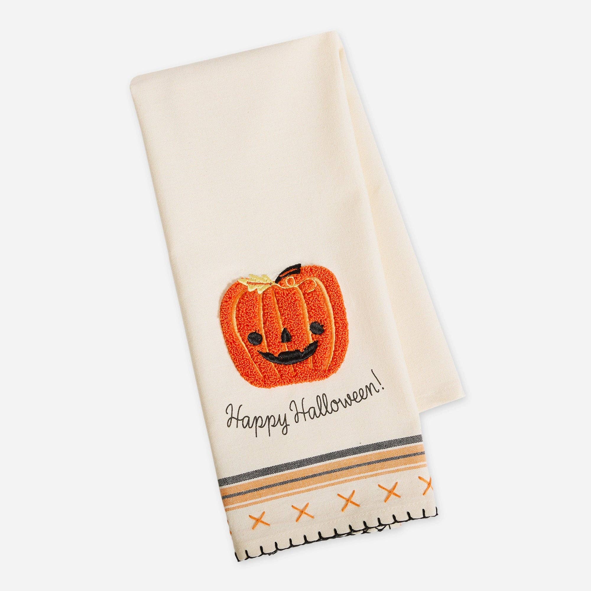 An off-white Halloween kitchen towel featuring an embroidered orange jack-o'-lantern with a smiling face and the words 'Happy Halloween!' in black script. The bottom edge has a decorative border with orange 'X' patterns and black stitched fringe.