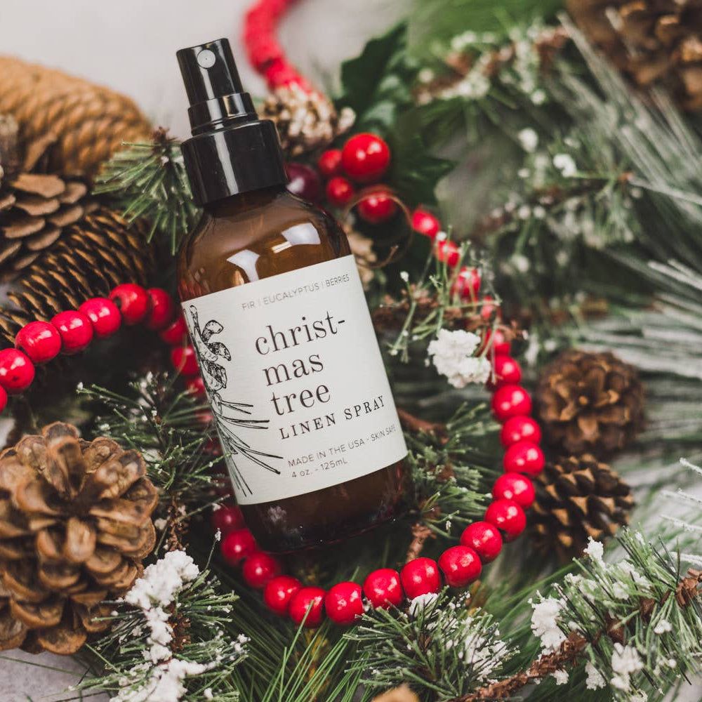 Bottle of Christmas tree linen spray surrounded by festive decorations including pine cones and red berries.