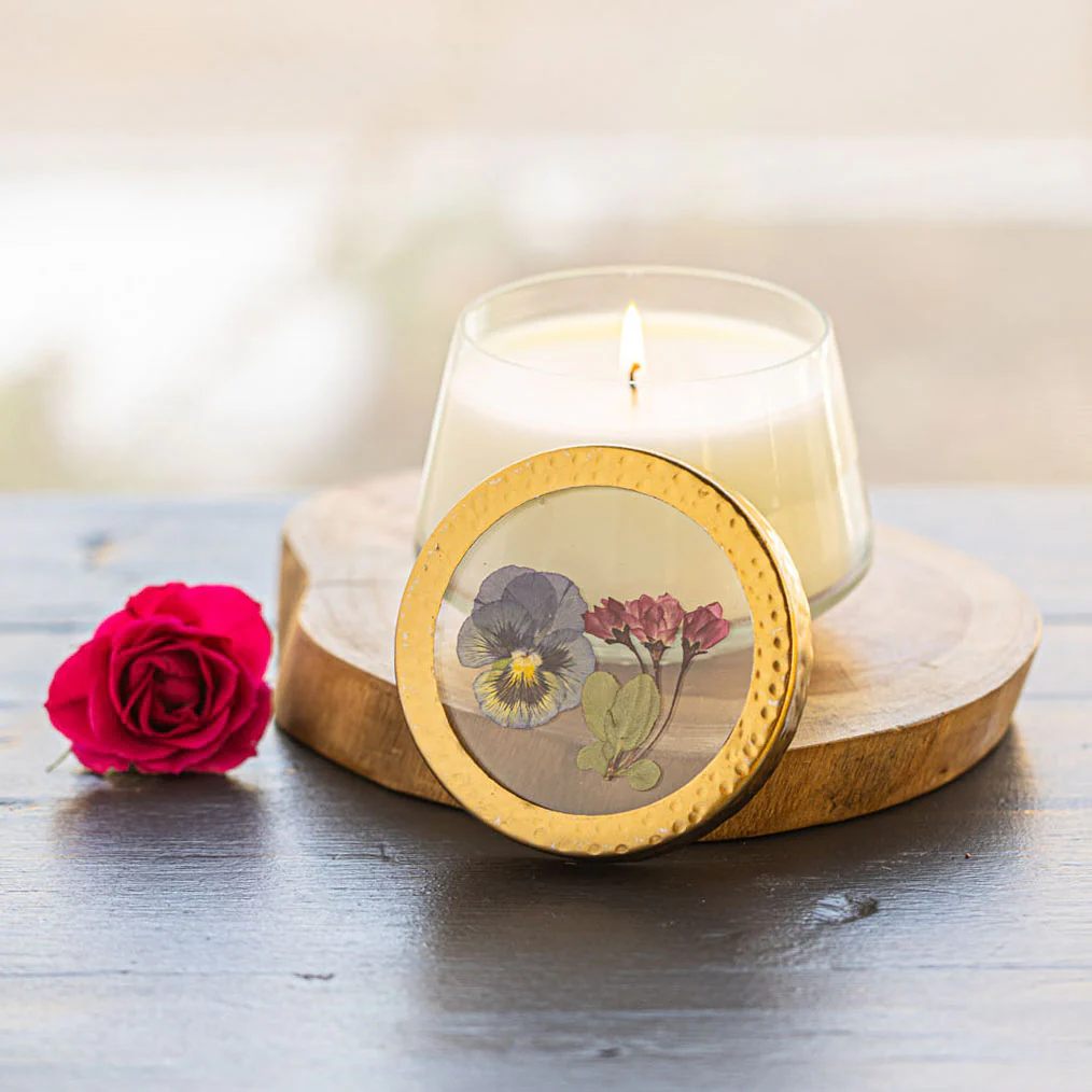 Candle in a glass holder with floral design on a wooden coaster, next to a pink rose.
