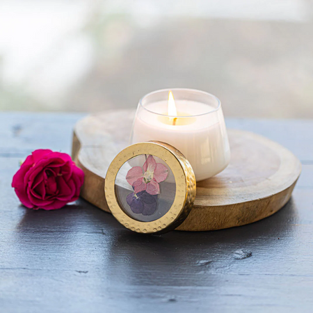 Candle with a decorative lid featuring pressed flowers, next to a pink rose on a wooden coaster.