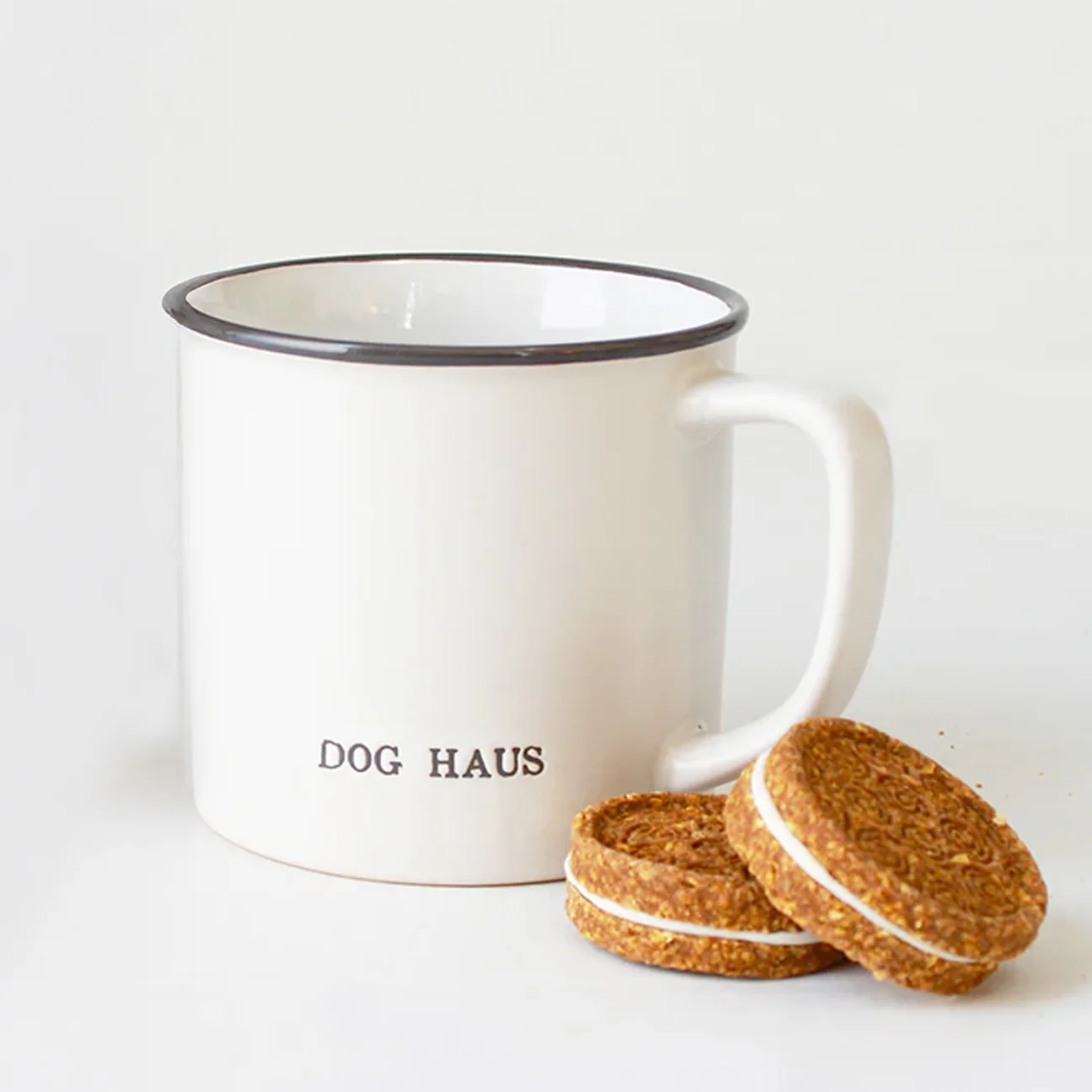 A white enamel-style mug with a black rim and the words 'DOG HAUS' printed on the front, shown on a white background with two sandwich cookies placed in front.