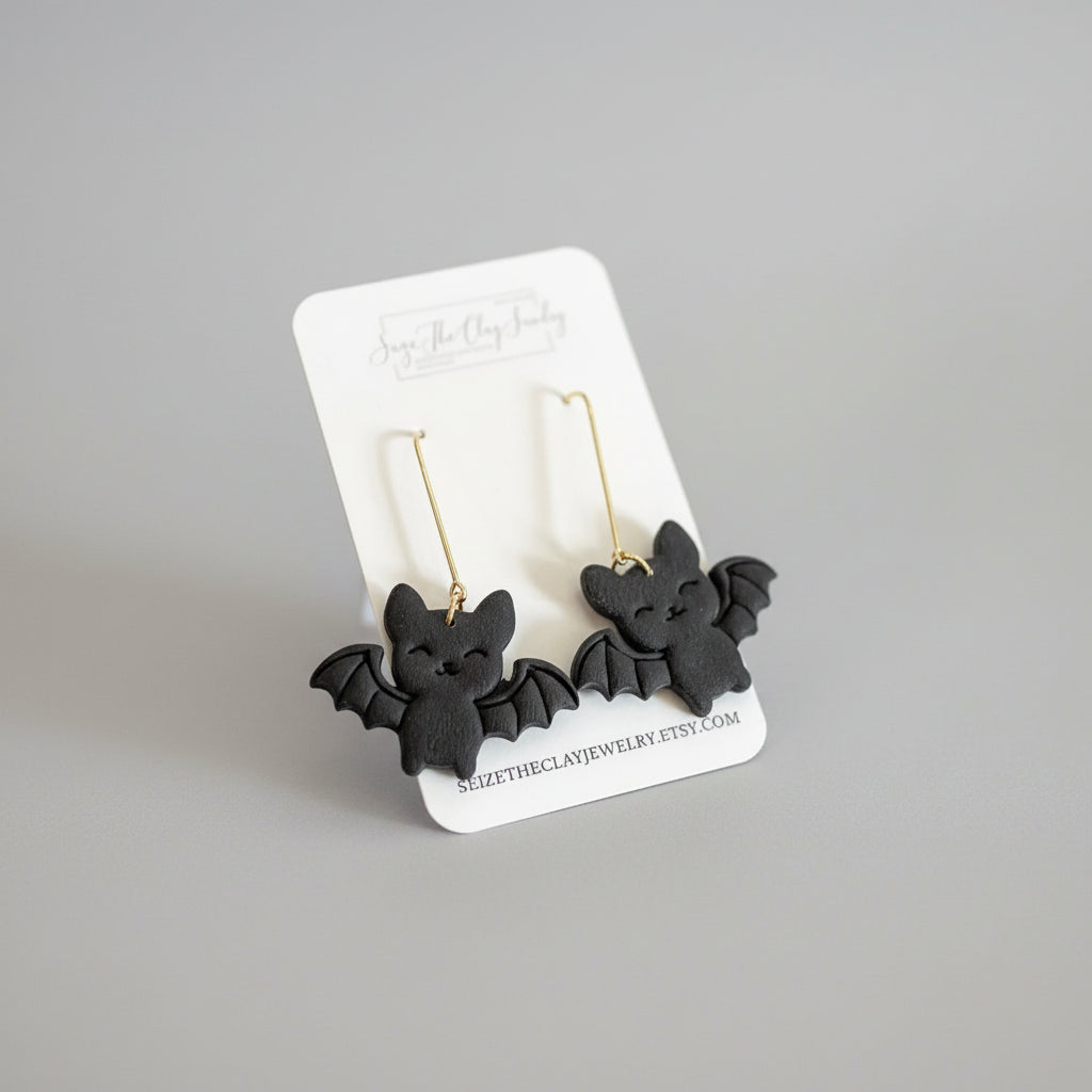 A pair of black bat-shaped drop earrings with gold hooks, displayed on a white branded card against a textured magenta background.