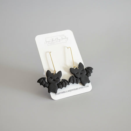 A pair of black bat-shaped drop earrings with gold hooks, displayed on a white branded card against a textured magenta background.