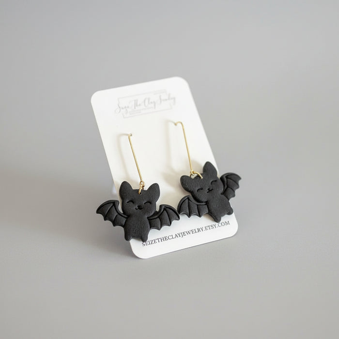 A pair of black bat-shaped drop earrings with gold hooks, displayed on a white branded card against a textured magenta background.