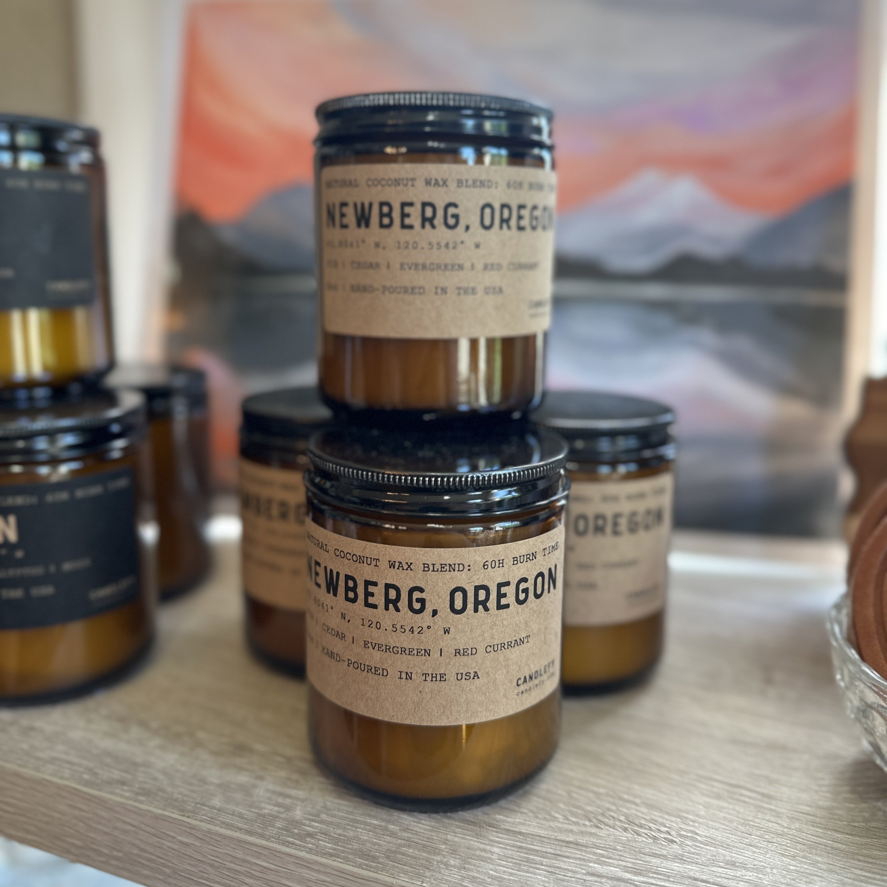 Amber glass Newberg, Oregon candle with labels on a wooden shelf, with a scenic background.